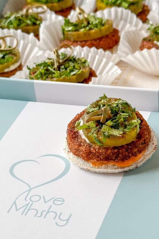 Small appetizers with a green topping on a white paper, placed on a light blue surface with 'Love Mshy' branding.